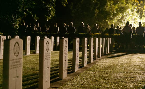 War graves at a military cemetery with visitors gathered in the background during a commemorative event.